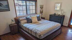 A bedroom with a large bed, wooden headboard, a nightstand, a dresser, and a window with a view of a wooden fence.