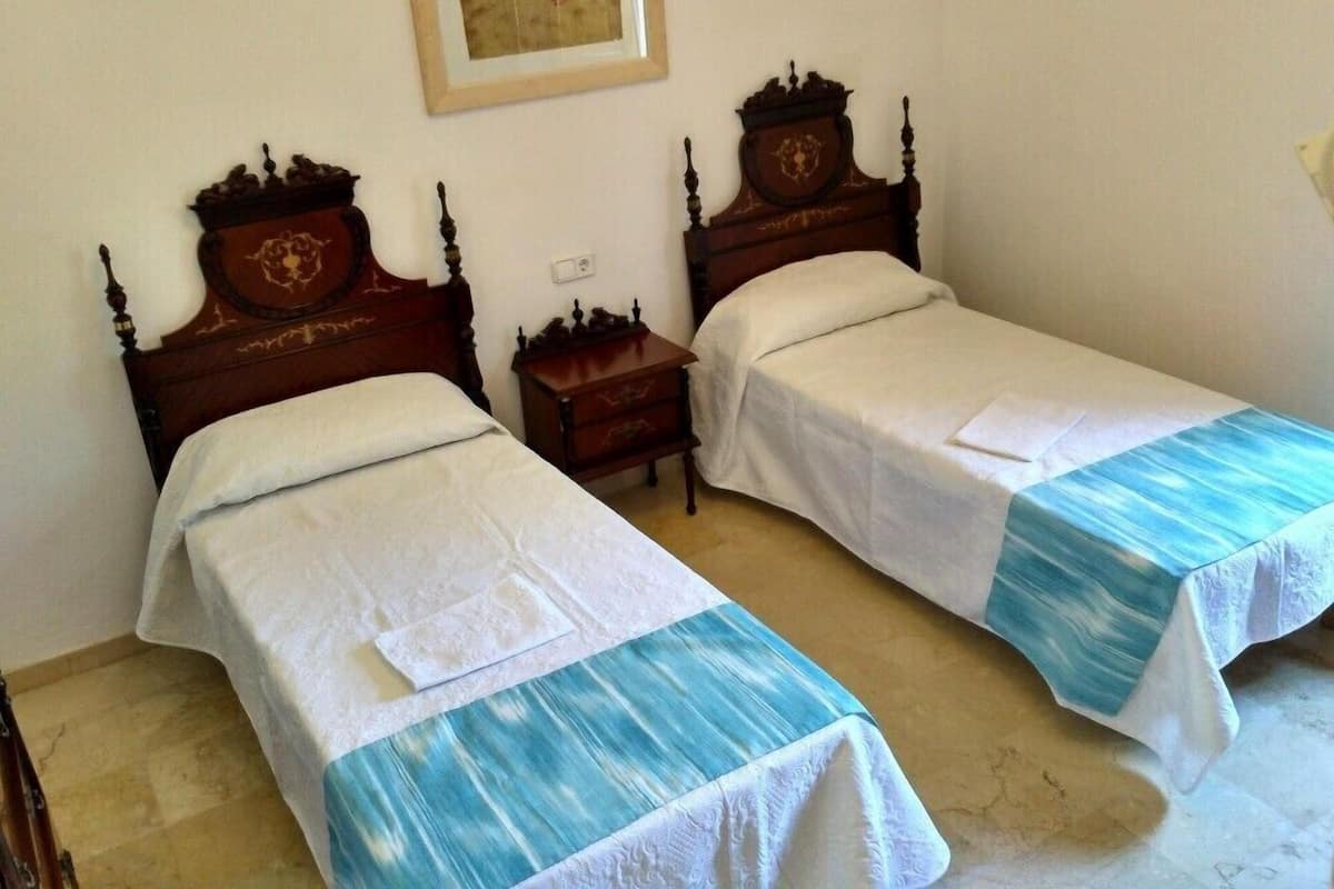 Ein Zimmer mit zwei Holzbetten, jeweils mit weißer Bettdecke und einer blau-weißen Streifenwolldecke. Zwischen den Betten steht ein kleiner Nachttisch aus Holz, darüber an der Wand ein gerahmtes Bild.