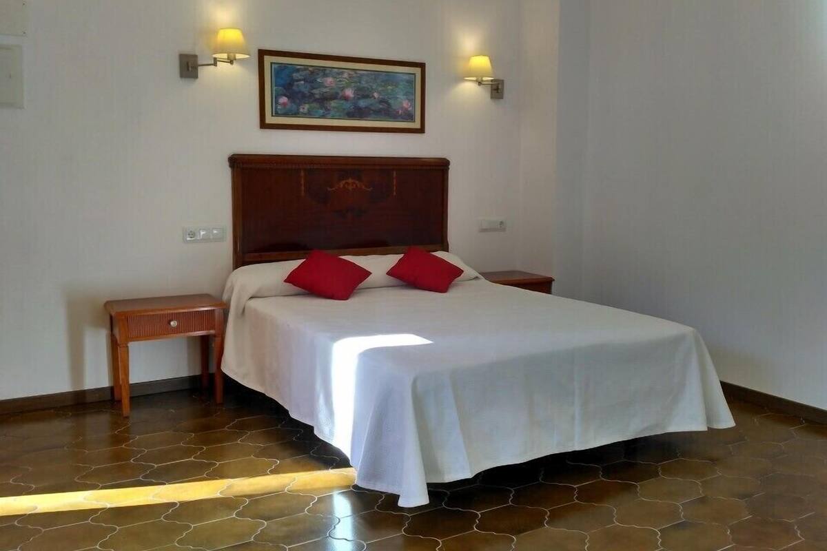 Ein Schlafzimmer mit einem hölzernen Kopfende, einer weißen Bettdecke, zwei roten Kissen, einem Nachttisch und einem Bild an der Wand.
