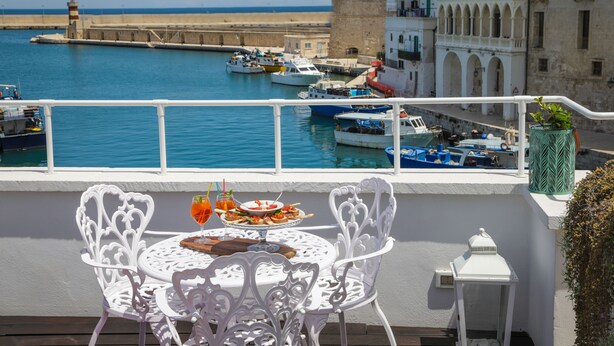 A balcony with white ornate chairs and a table set for dining, overlooking a harbor with boats and a historic building. - Al Console Luxury Suite (Monopoli)
