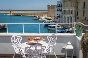 A balcony with white ornate chairs and a table set for dining, overlooking a harbor with boats and a historic building. - Al Console Luxury Suite (Monopoli)
