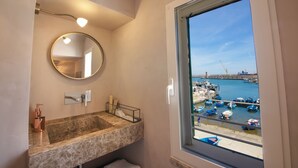 Un baño con lavamanos de mármol, un espejo redondo y una ventana con vistas a un puerto deportivo con barcos.