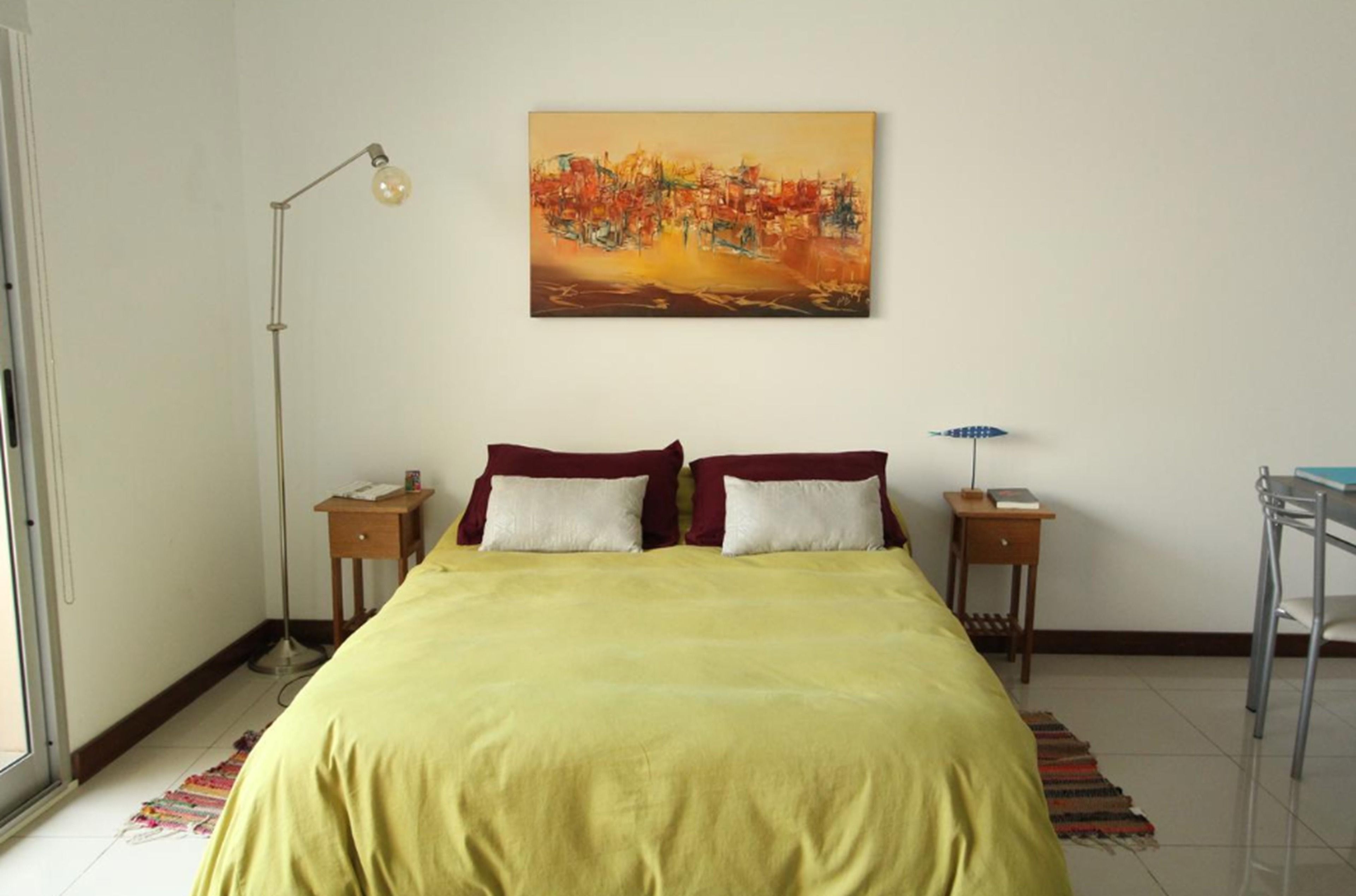 A bedroom with a bed, a painting on the wall, a floor lamp, and a desk with a chair.