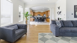 A modern living room with a grey sofa, a grey armchair, and a patterned rug. A kitchen with wooden cabinets and a dining area with a table and chairs is visible in the background. - Vilano Beach Pool Home-Ocean Views (St. Augustine)
