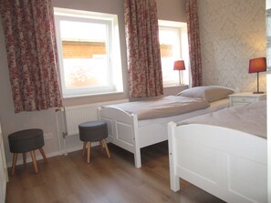 A bedroom with two single beds, a window with curtains, a radiator, a bedside table with a lamp, and a small stool. - Apartment Bendixen - Apartment Bendixen (Steinberg)