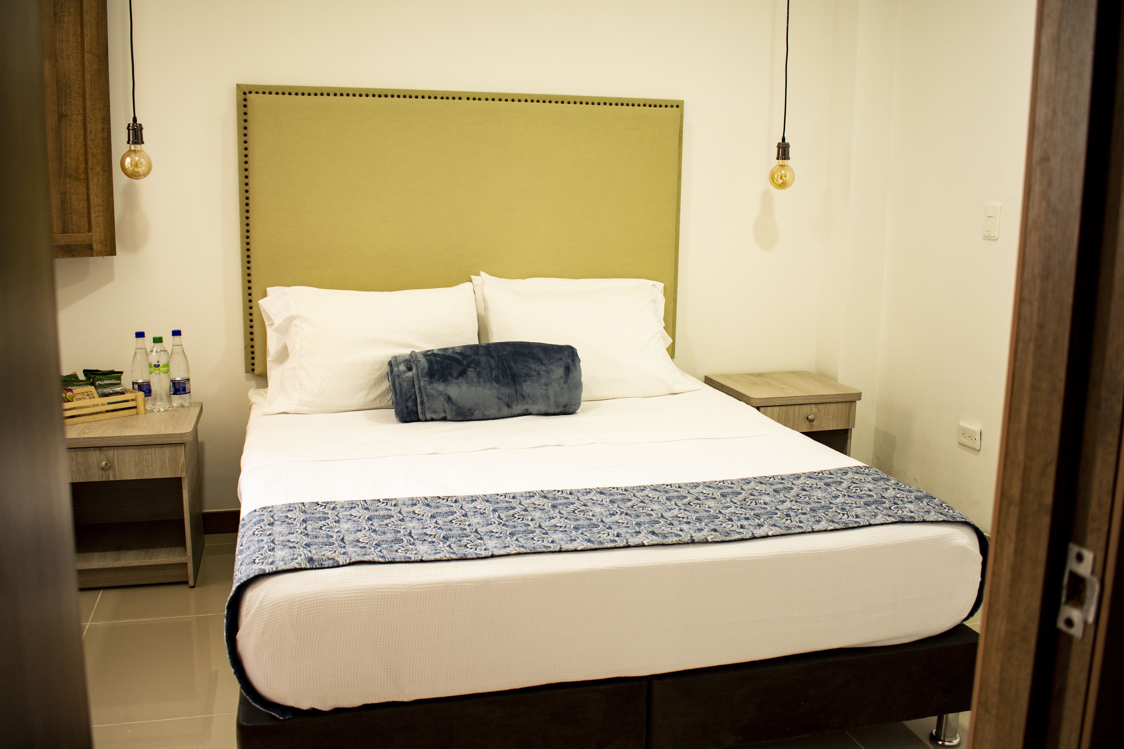 A bed with a headboard, pillows, and a blanket. There is a nightstand with bottles and a lamp.