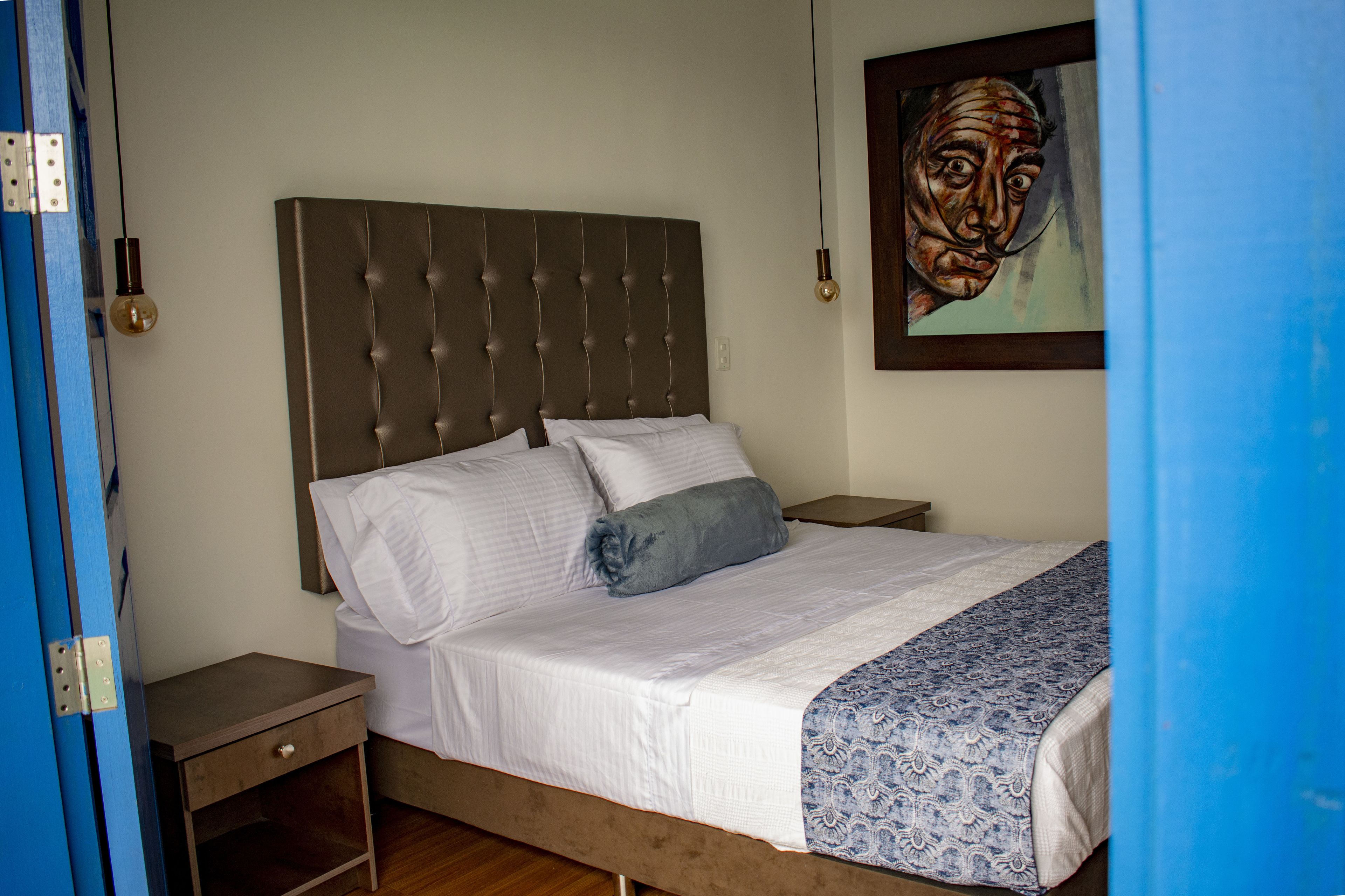 A bedroom with a tufted headboard, a bed with white linens, a patterned throw blanket, a nightstand, and a painting on the wall.