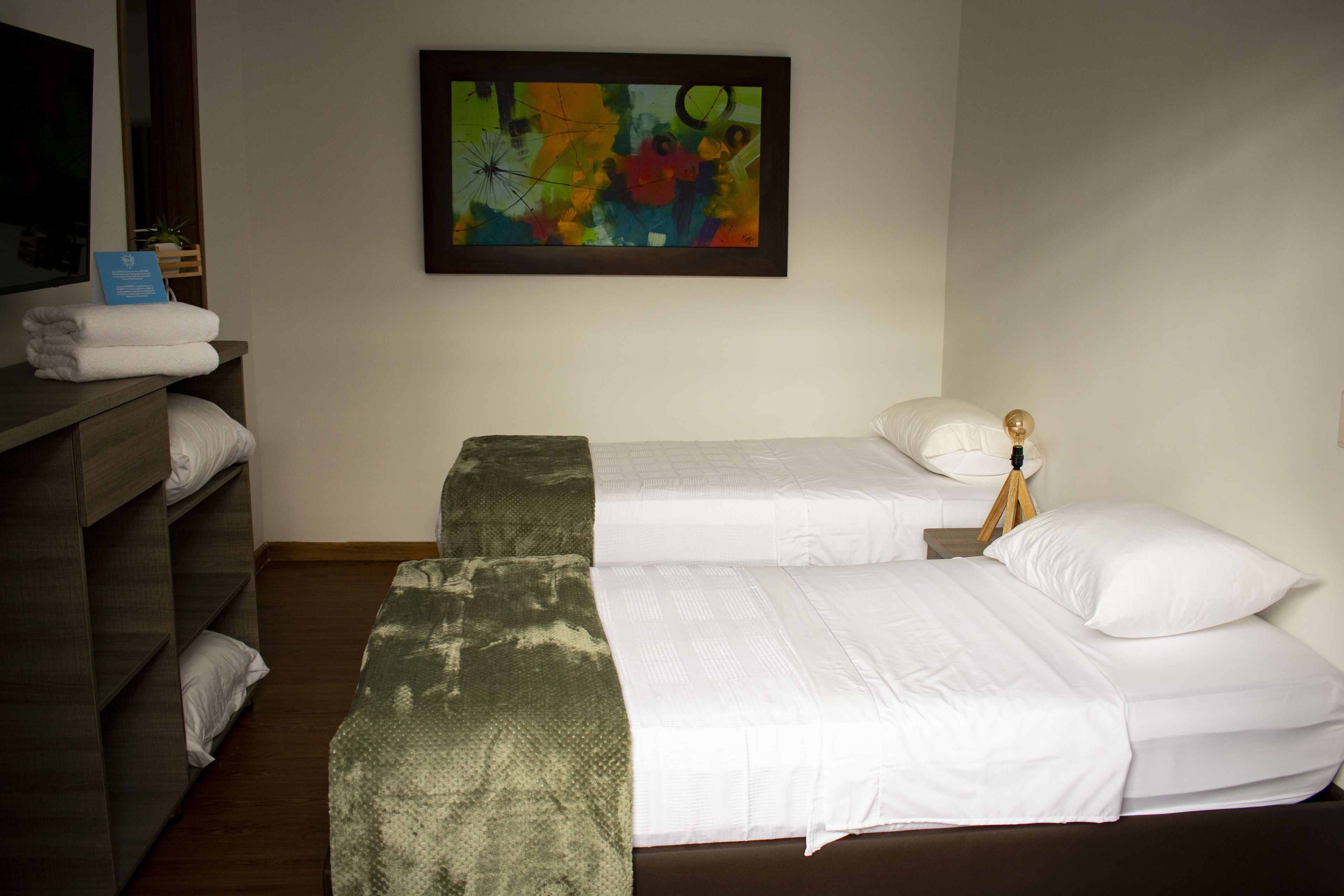 Quarto de hotel com duas camas, um aparador de madeira, uma pintura abstrata colorida e uma pequena planta.