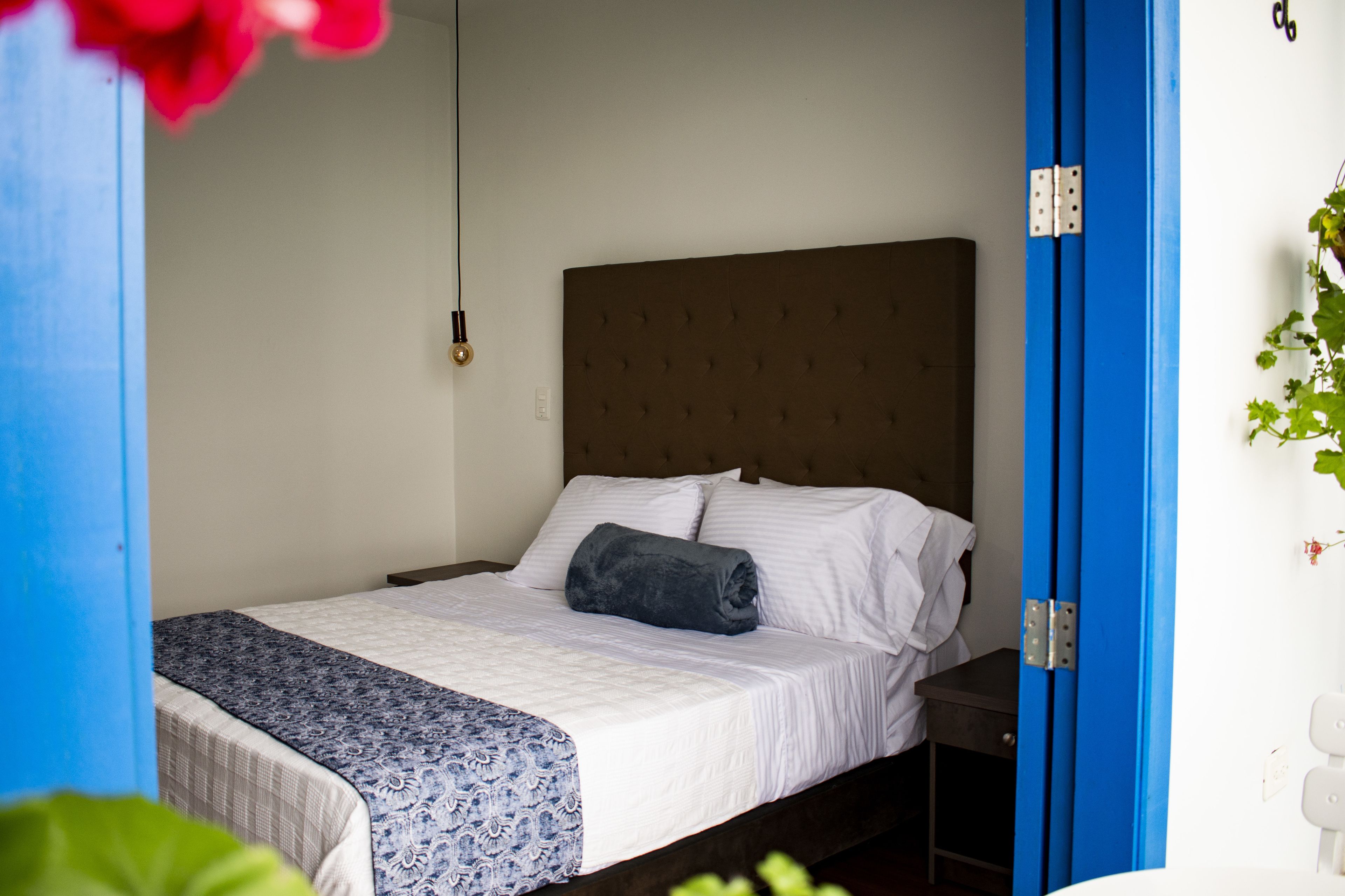 A neatly made bed with a patterned bedspread and pillows, a bedside table, and a blue door frame.