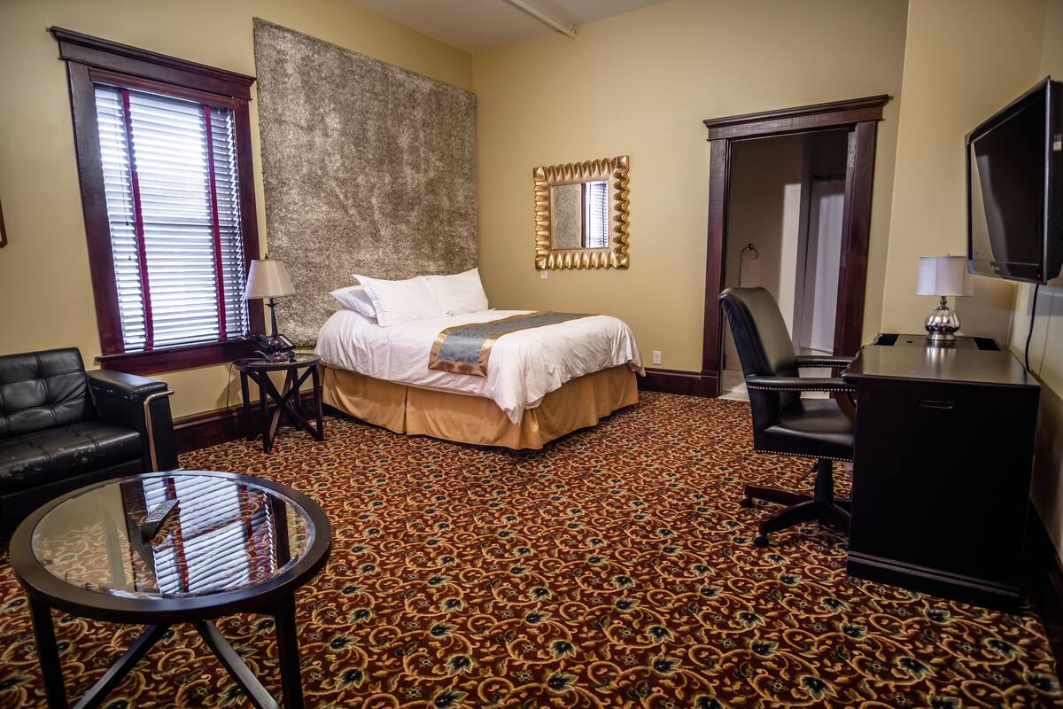 A hotel room with a large bed, a desk, a chair, a round table, and a large window with blinds.