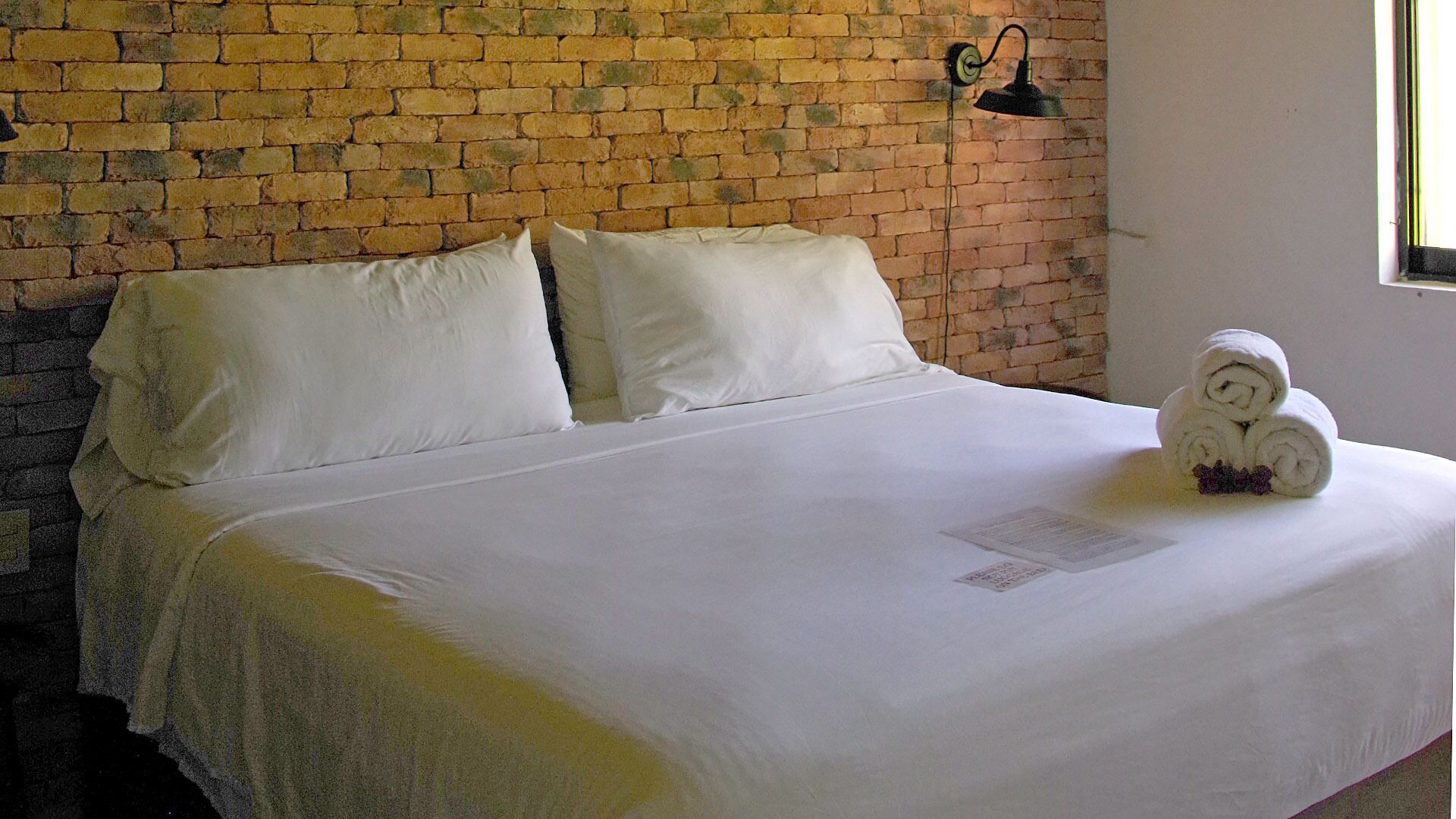 A neatly made bed with white linens, a brick wall background, and a small towel arrangement on the bed.