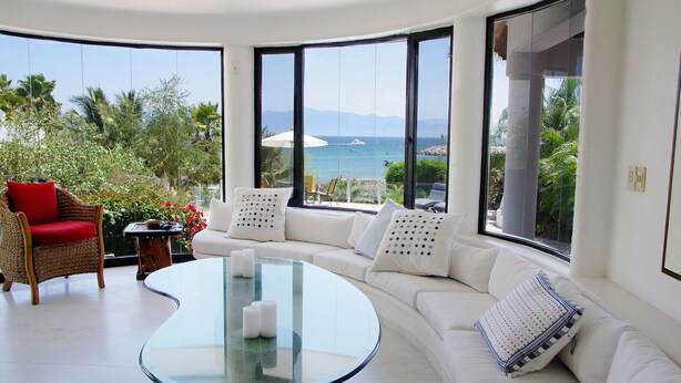 Un salon moderne avec une table basse en verre, un canapé blanc et un fauteuil en osier agrémenté de coussins rouges. De grandes fenêtres offrent une vue sur la mer et les palmiers.