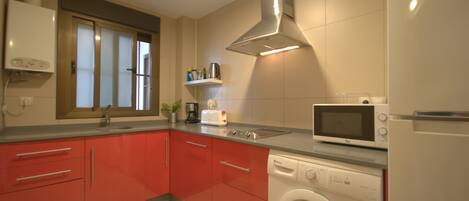 A modern kitchen with red cabinets, a microwave, a washing machine, and a window with blinds.