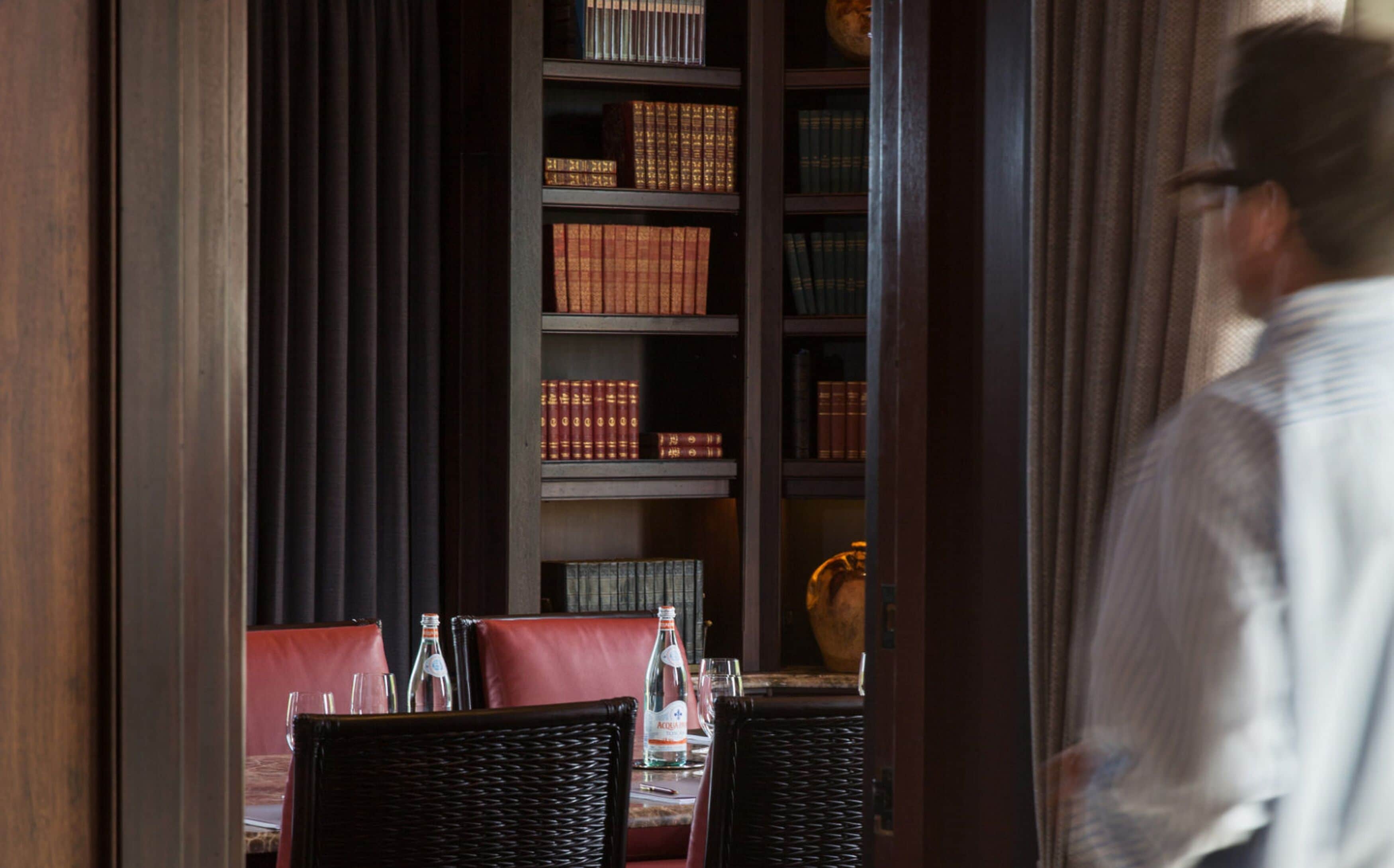 A restaurant interior with a dark wood bookshelf filled with books, leather chairs, and a table set for dining.