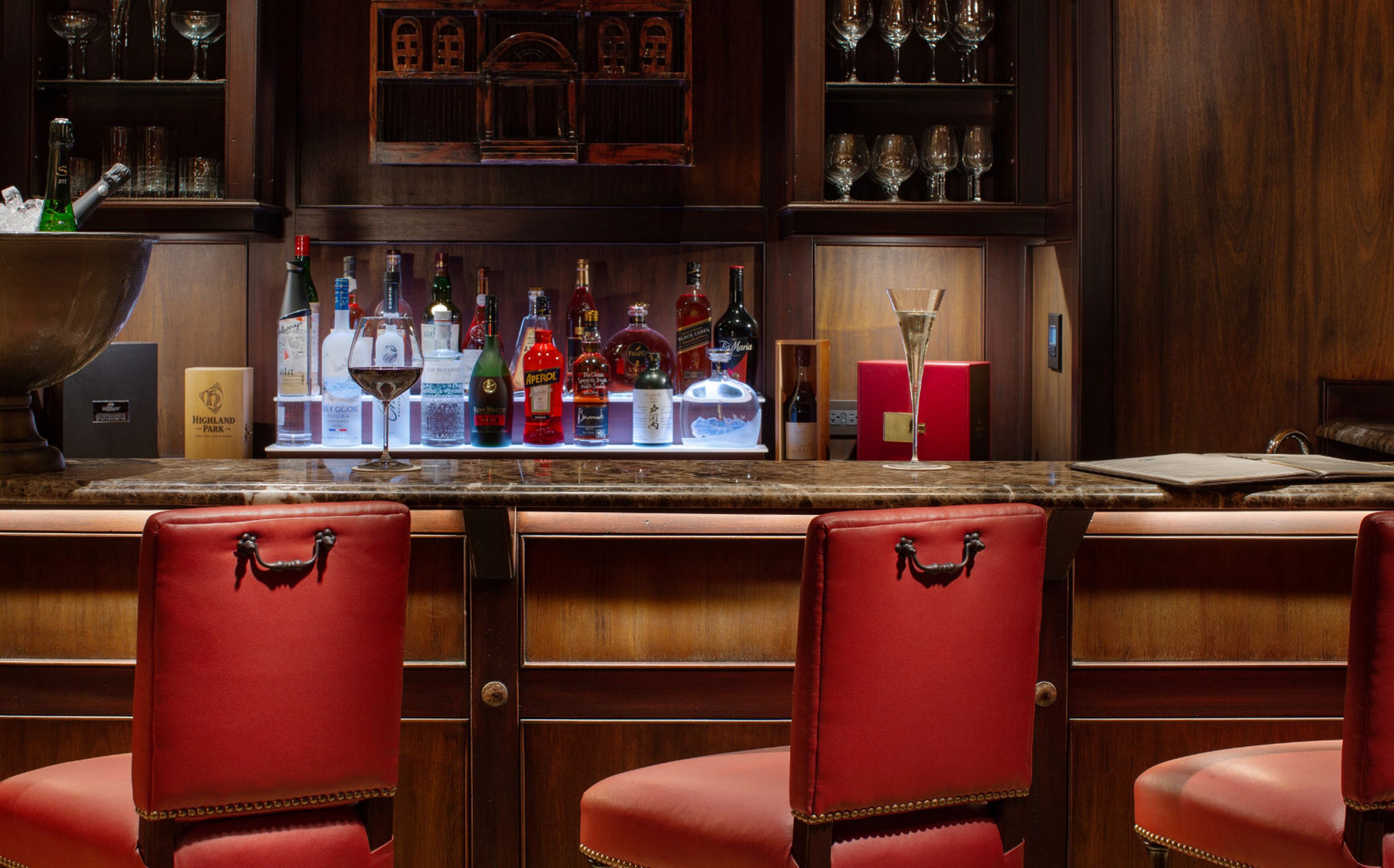 Una zona de bar con mostrador de madera, varias botellas de licores y taburetes de cuero rojo.