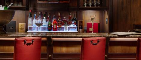 Una zona de bar con mostrador de madera, varias botellas de licores y taburetes de cuero rojo.