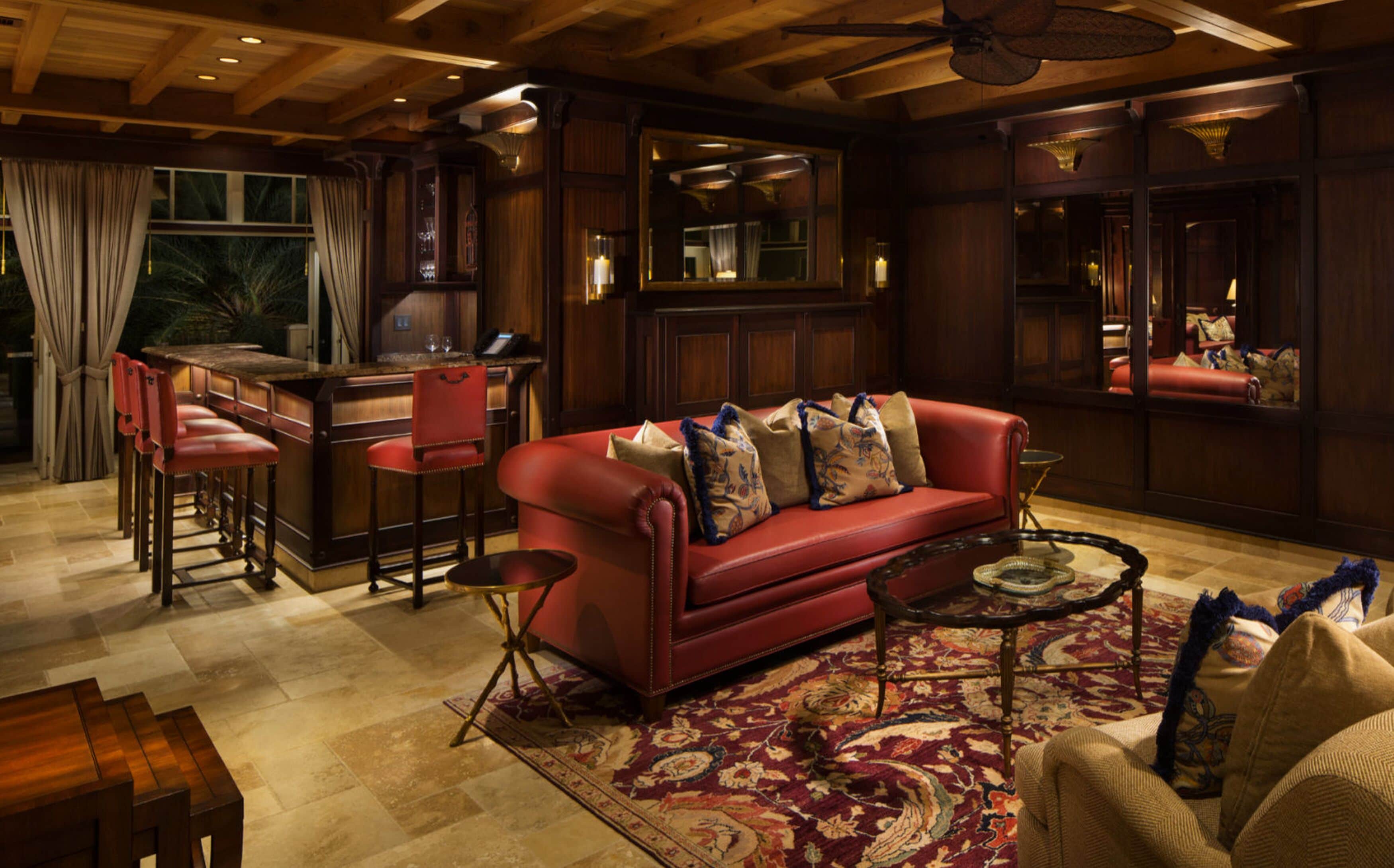 A cozy living room with a red leather sofa, a coffee table, and a patterned rug. There is a bar area with stools and a television.