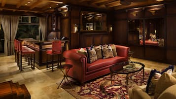 A cozy living room with a red leather sofa, a coffee table, and a patterned rug. There is a bar area with stools and a television.