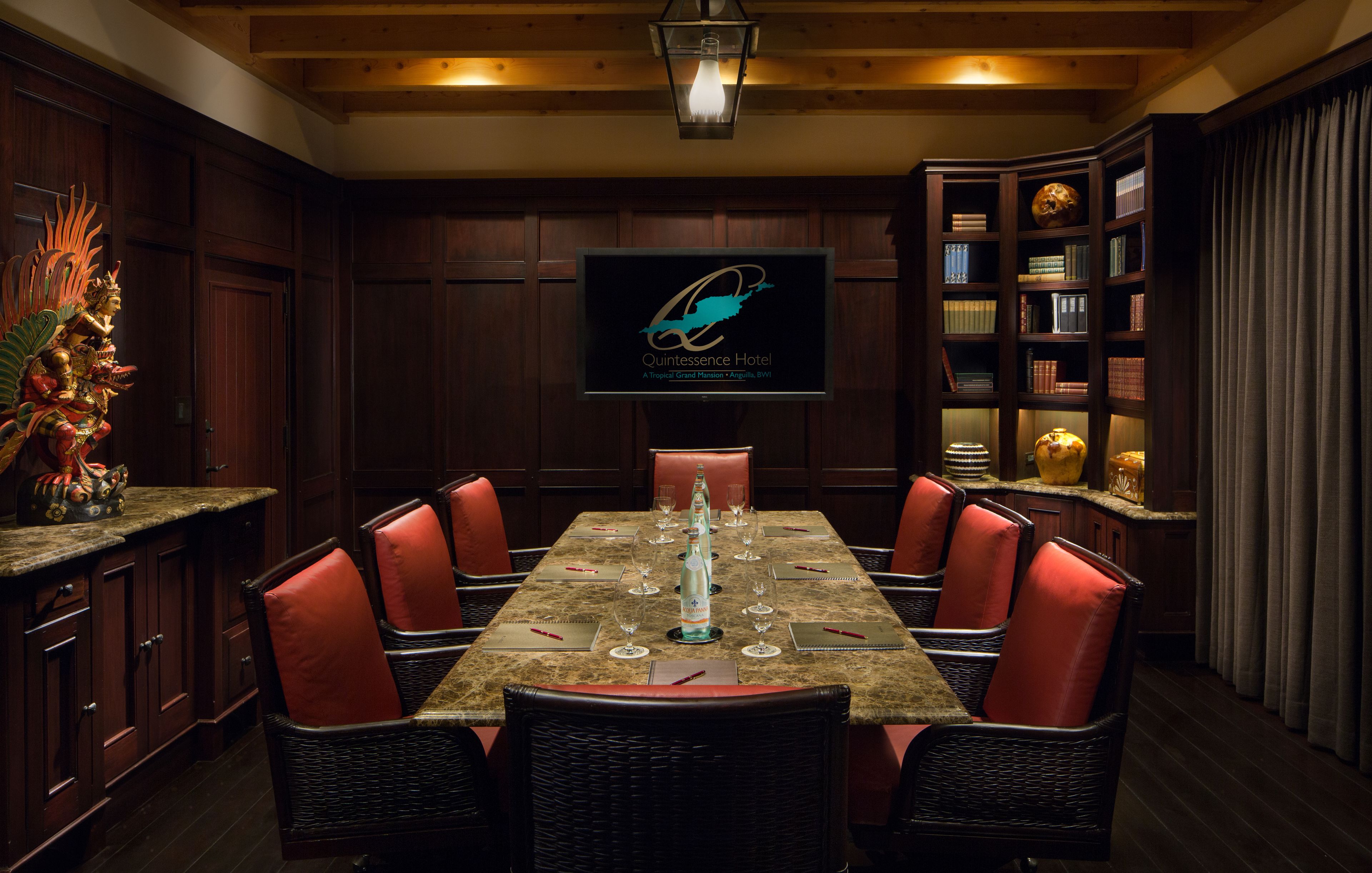 A dining area with a large table set for six, surrounded by wicker chairs with red cushions. A television screen displays the hotel logo.