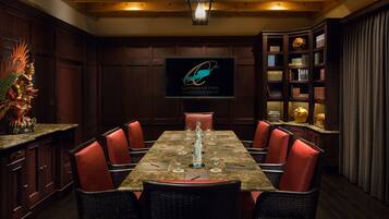 A dining area with a large table set for six, surrounded by wicker chairs with red cushions. A television screen displays the hotel logo.