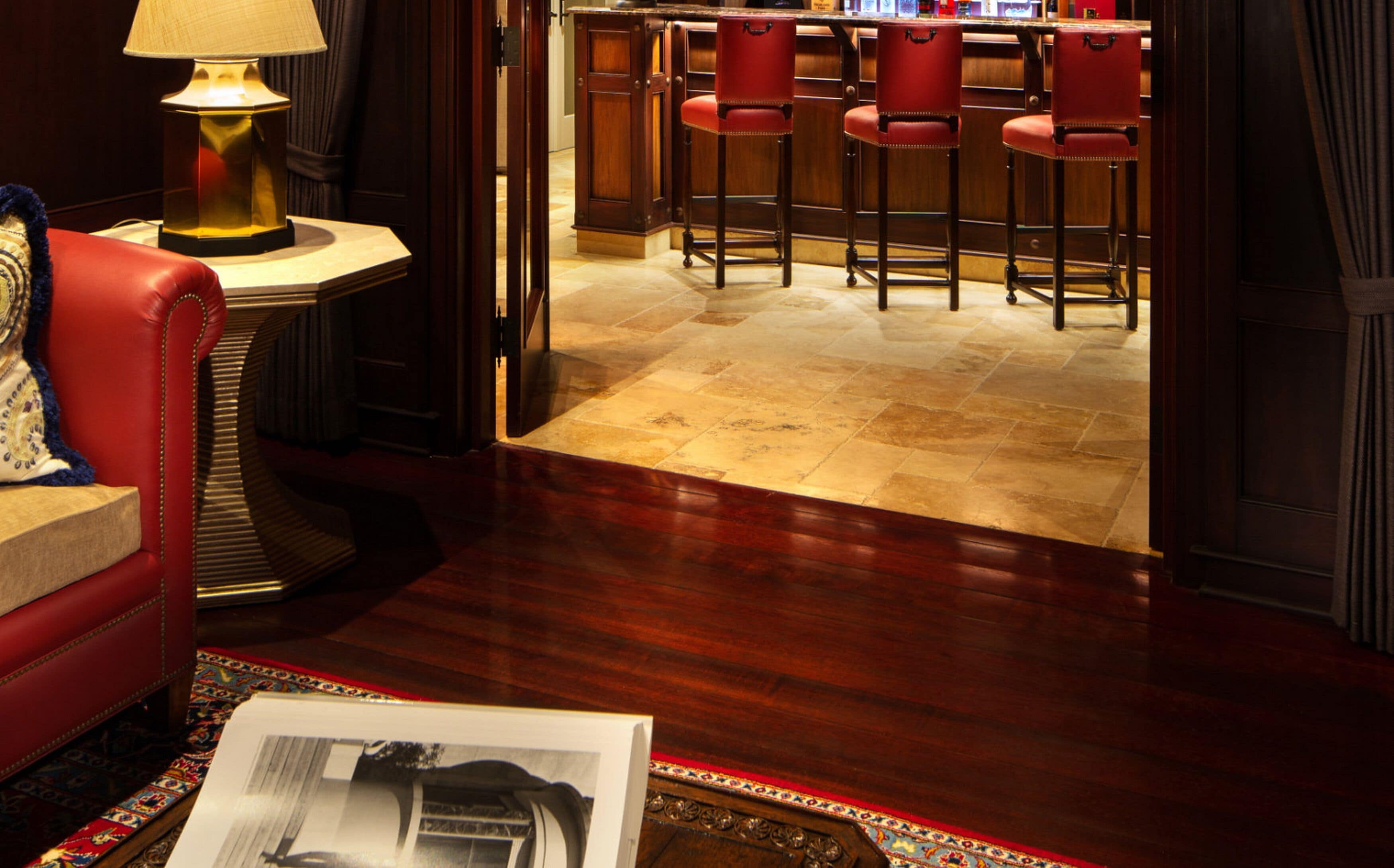 A hotel lobby with a red sofa, a table lamp, a bar area with red chairs, and a patterned rug.