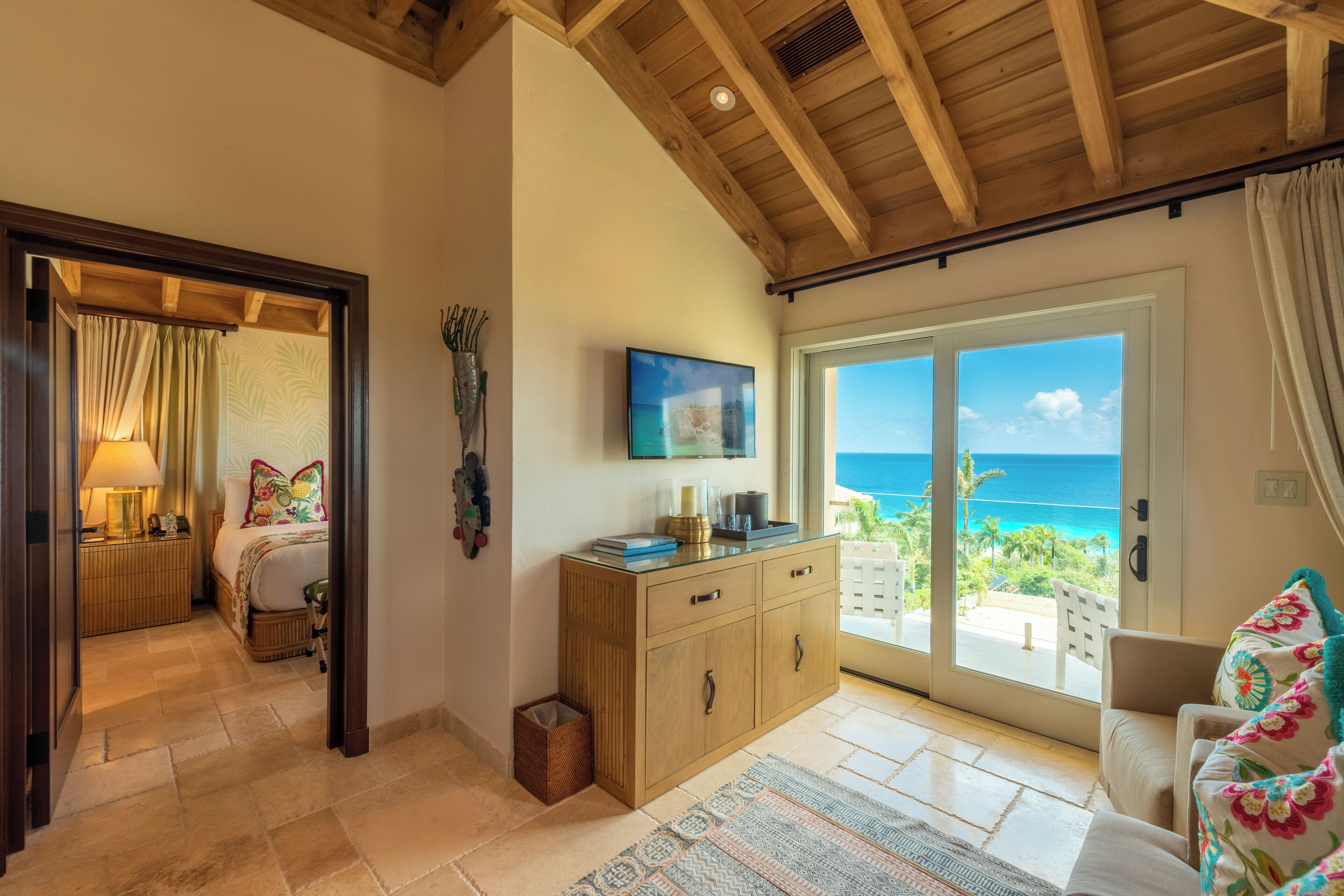 Un dormitorio con una cama, una mesita de noche, una lámpara, un aparador y un televisor. Hay una puerta corrediza de vidrio que da a un balcón con vista al océano.