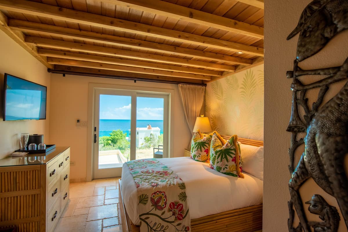 Ein Schlafzimmer mit Holzboden, einem Bett mit Blumenmuster-Bettwäsche, einem Fernseher auf der linken Seite und Blick auf das Meer durch die Tür.