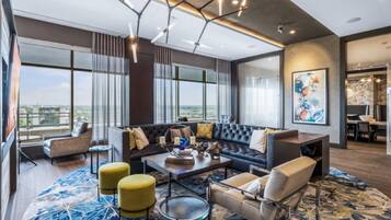 A modern living room with a large sofa, armchairs, and ottomans. There are large windows offering a view of the city.