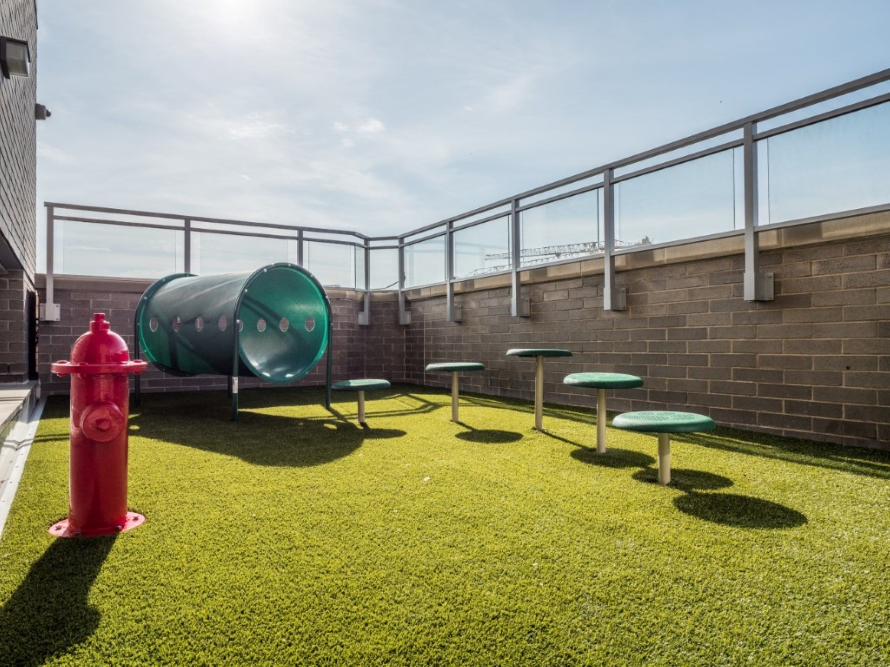 Zona de juegos al aire libre con césped artificial, un hidrante rojo, estructuras de juego cilíndricas verdes y mesas de metal.