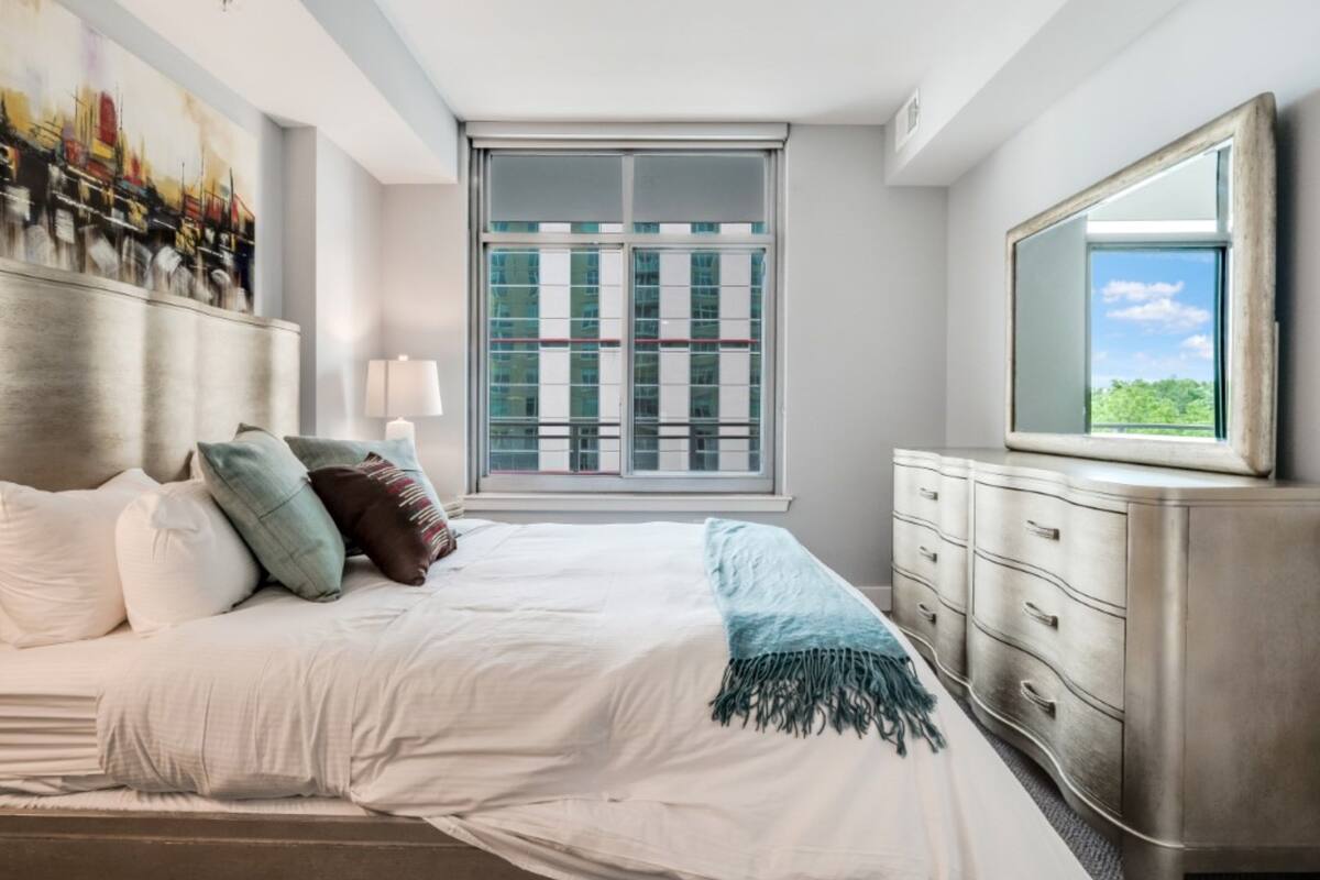 A modern bedroom with a large bed, a dresser, a painting on the wall, and a window with a view of buildings.