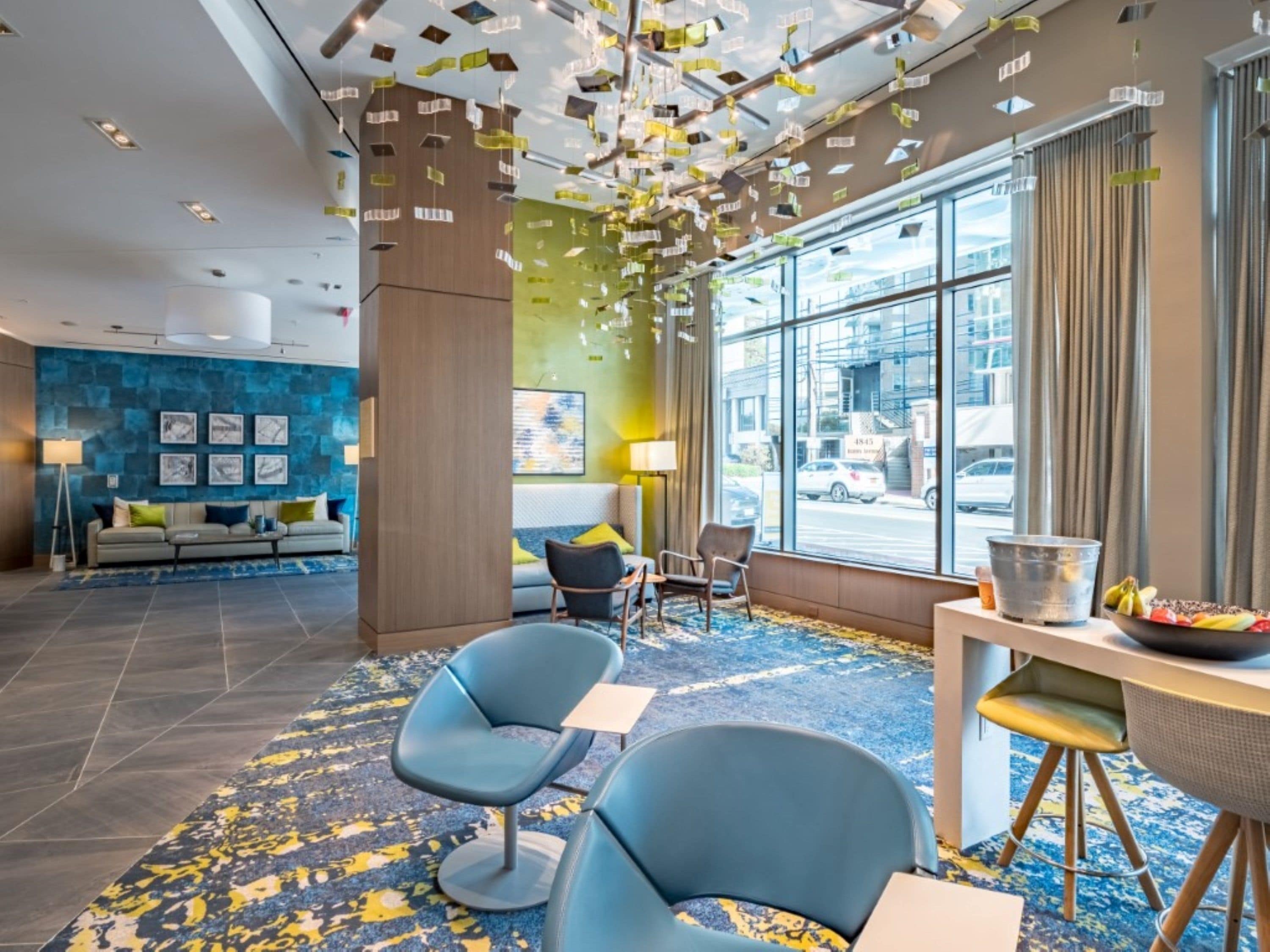 En modern hotellobby med ett blått och gult mönstrat golv, en sittgrupp med soffor och fåtöljer samt ett stort fönster med utsikt över staden.