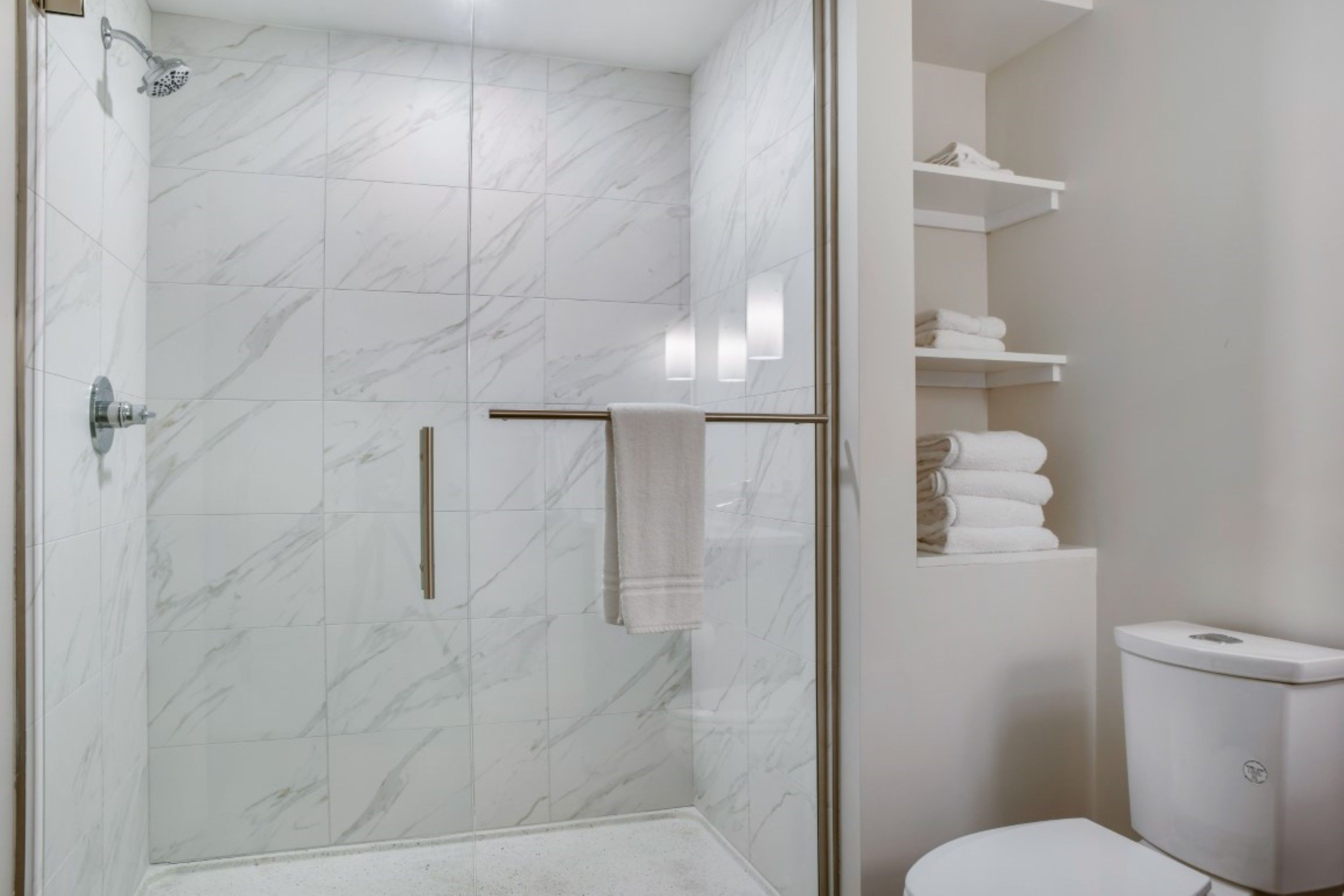 Une salle de bain moderne avec une douche à parois de verre, des carreaux de marbre, des toilettes et des étagères blanches où sont posées des serviettes.