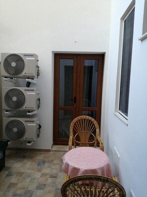 Une petite pièce avec un sol carrelé, une porte en bois et une table recouverte d’une nappe rose. Trois unités de climatisation sont fixées au mur.
