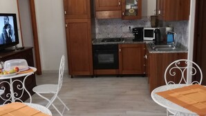 Une cuisine compacte avec des meubles en bois, un four à micro-ondes, une cuisinière et un évier. On y trouve deux chaises en métal blanches et une petite table avec un plateau en bois. Un téléviseur est fixé au mur.