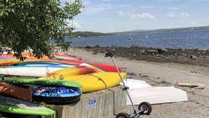 Eine Reihe bunter Kajaks und ein weißes Raft liegen auf einem felsigen Strand, mit Blick auf Wasser und Schiffe in der Ferne.
