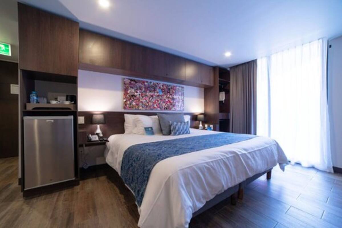 A modern hotel room with a large bed, a built-in refrigerator, a bedside table with a lamp, and a large window with curtains.