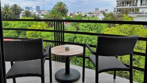 Un balcón con dos sillones de mimbre y una pequeña mesa redonda, con vistas a un exuberante jardín y edificios de la ciudad.