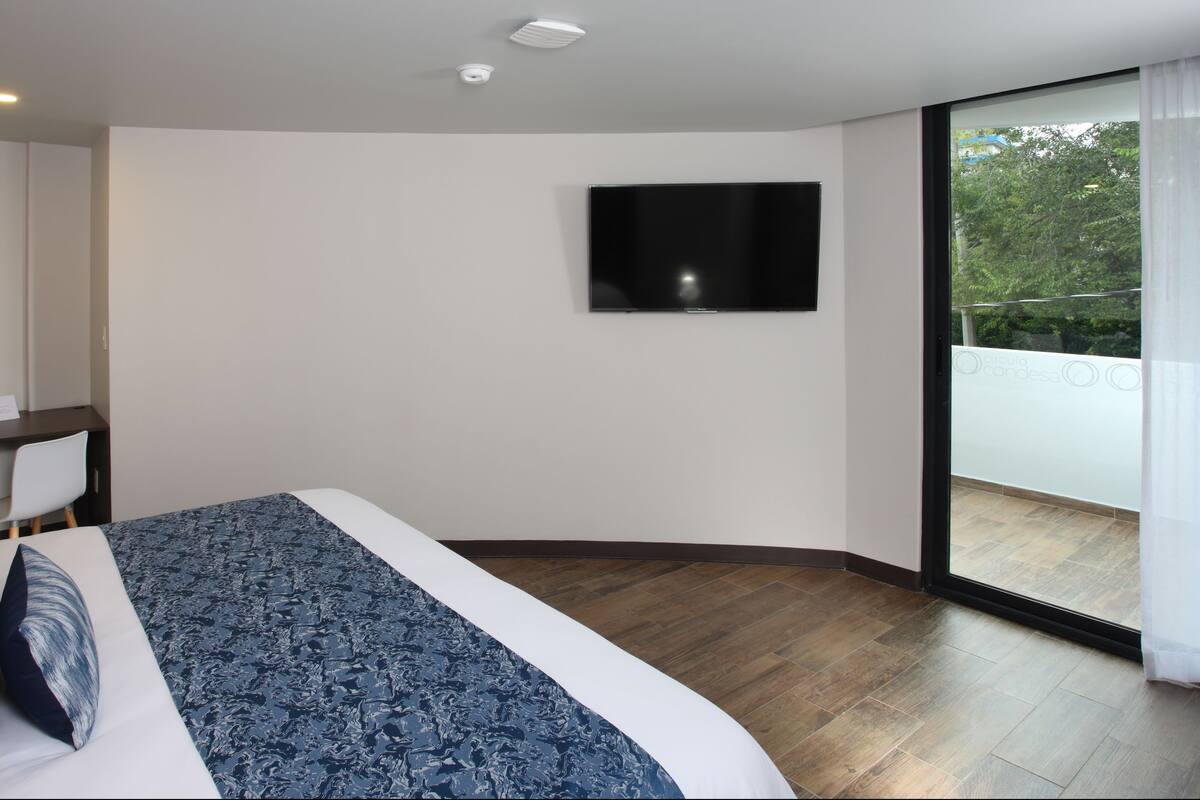 A modern hotel room with a large bed, a desk, a chair, a flat-screen TV mounted on the wall, and a sliding glass door leading to an outdoor balcony.