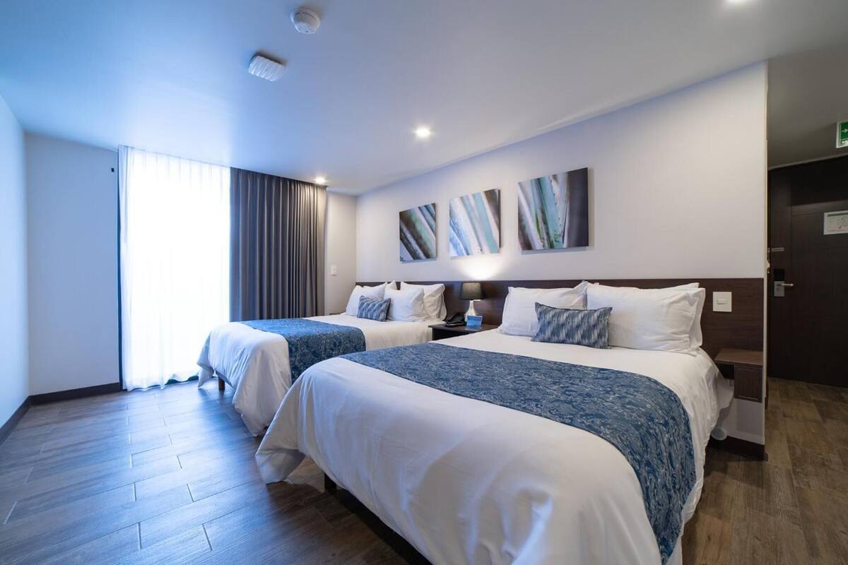 A hotel room with two beds, a large window with curtains, a wall-mounted artwork, and a wooden floor.
