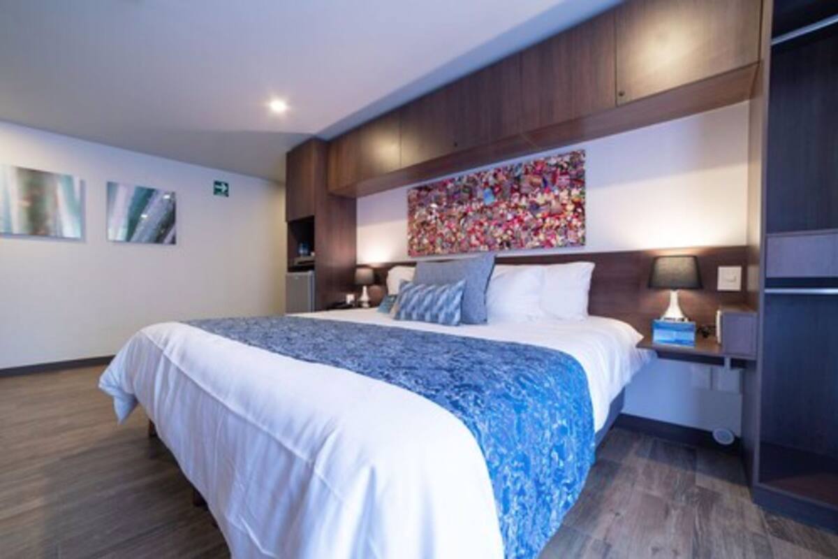 A hotel room with a large bed, wooden headboard, bedside tables with lamps, and a colorful artwork above the bed.