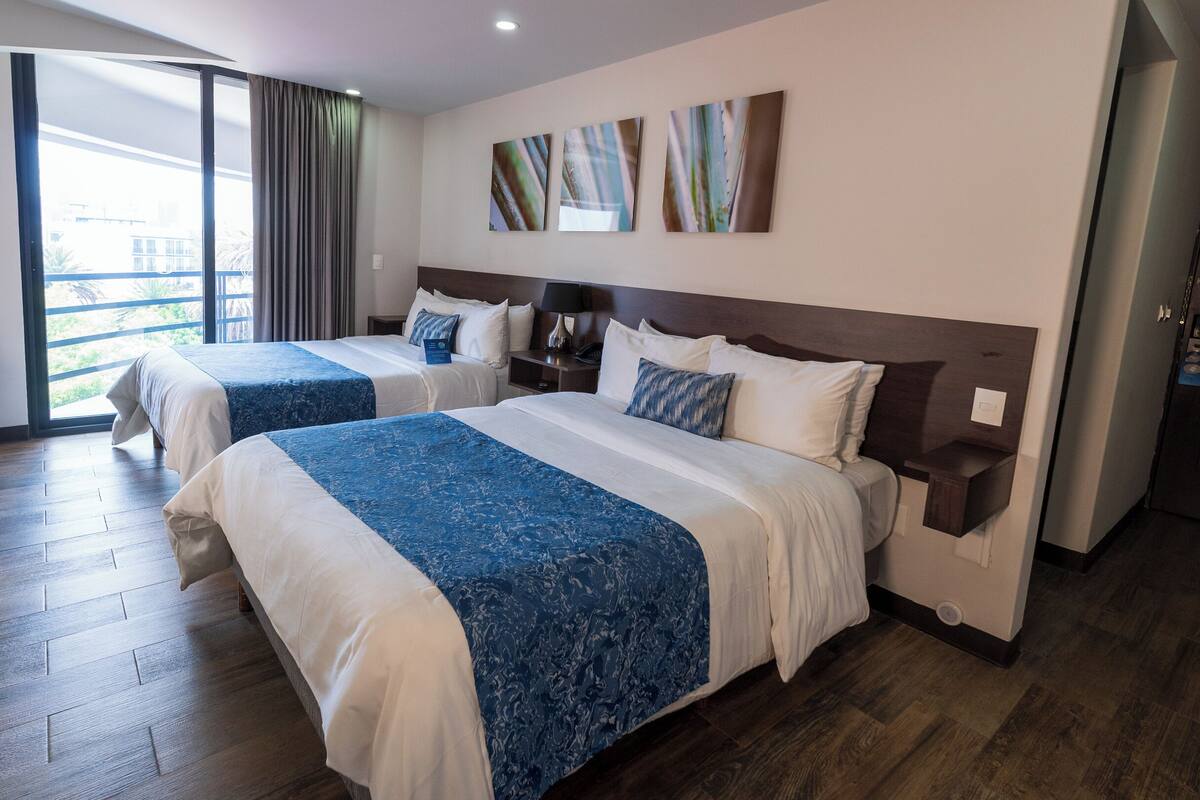 A hotel room with two beds, wooden flooring, a large window with curtains, and artwork on the wall.