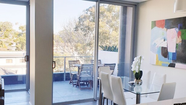 A modern kitchen with a glass dining table, white chairs, and a view of the outdoors through large sliding glass doors. - Modern 2b2b apartmemt. close to water front (Point Frederick)