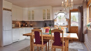 Una cocina con armarios blancos, una mesa de comedor de madera y sillas, una araña y un ventanal con cortinas.