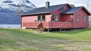 Una casa de madera roja con balcón, chimenea y vista a un lago y montañas al fondo.