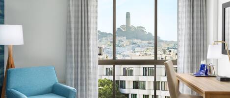 A hotel room with a large window offering a city view, a blue armchair, a wooden desk, and a small ottoman.