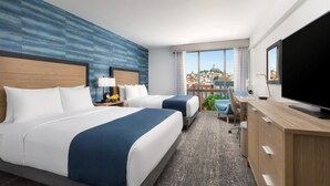 A hotel room with two beds, a desk, a TV, and a view of a cityscape.