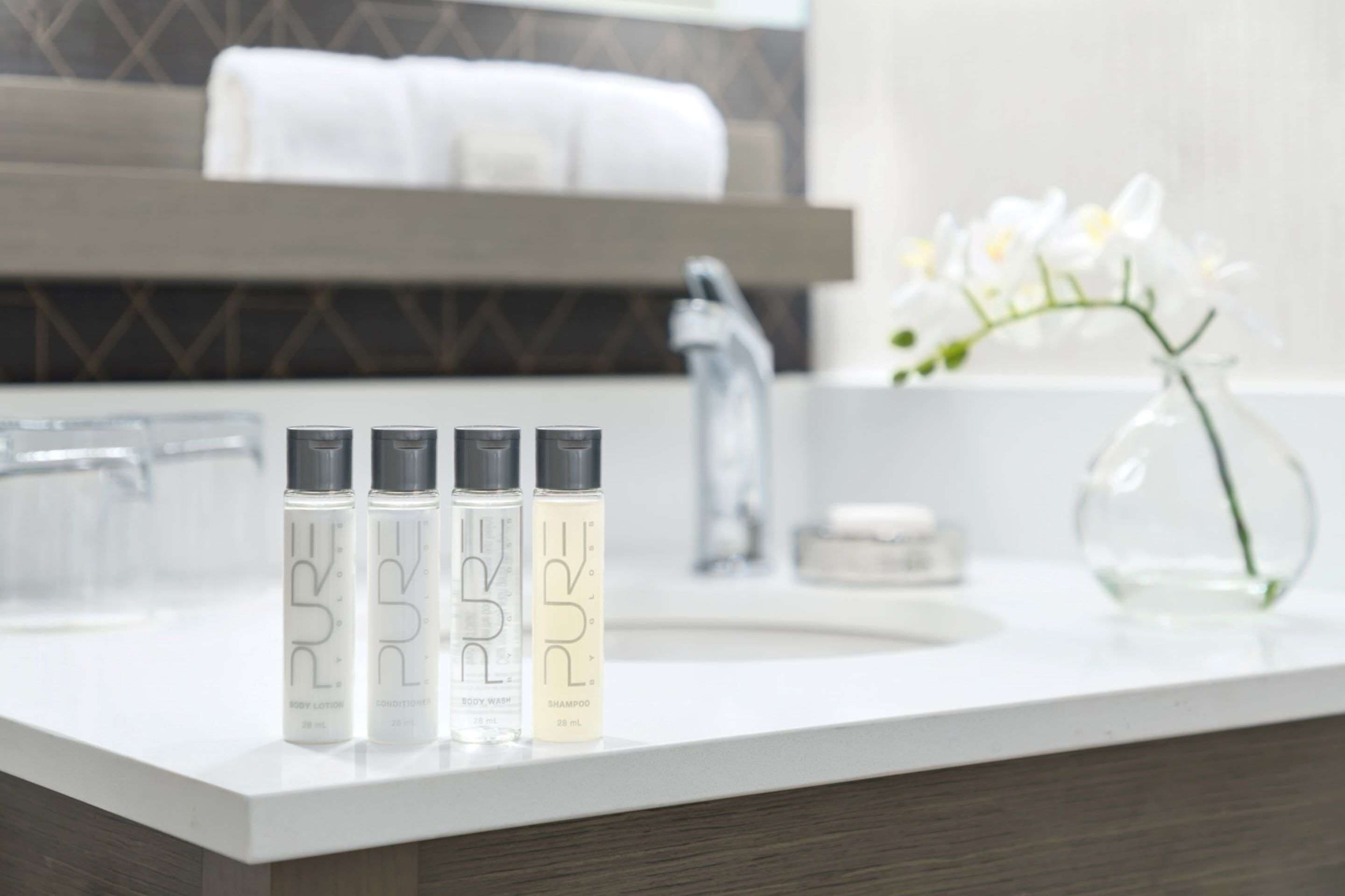 Un baño con un lavamanos blanco, tres botellas de productos corporales 'PURE', un florero con flores blancas y una toalla colgada en un perchero.