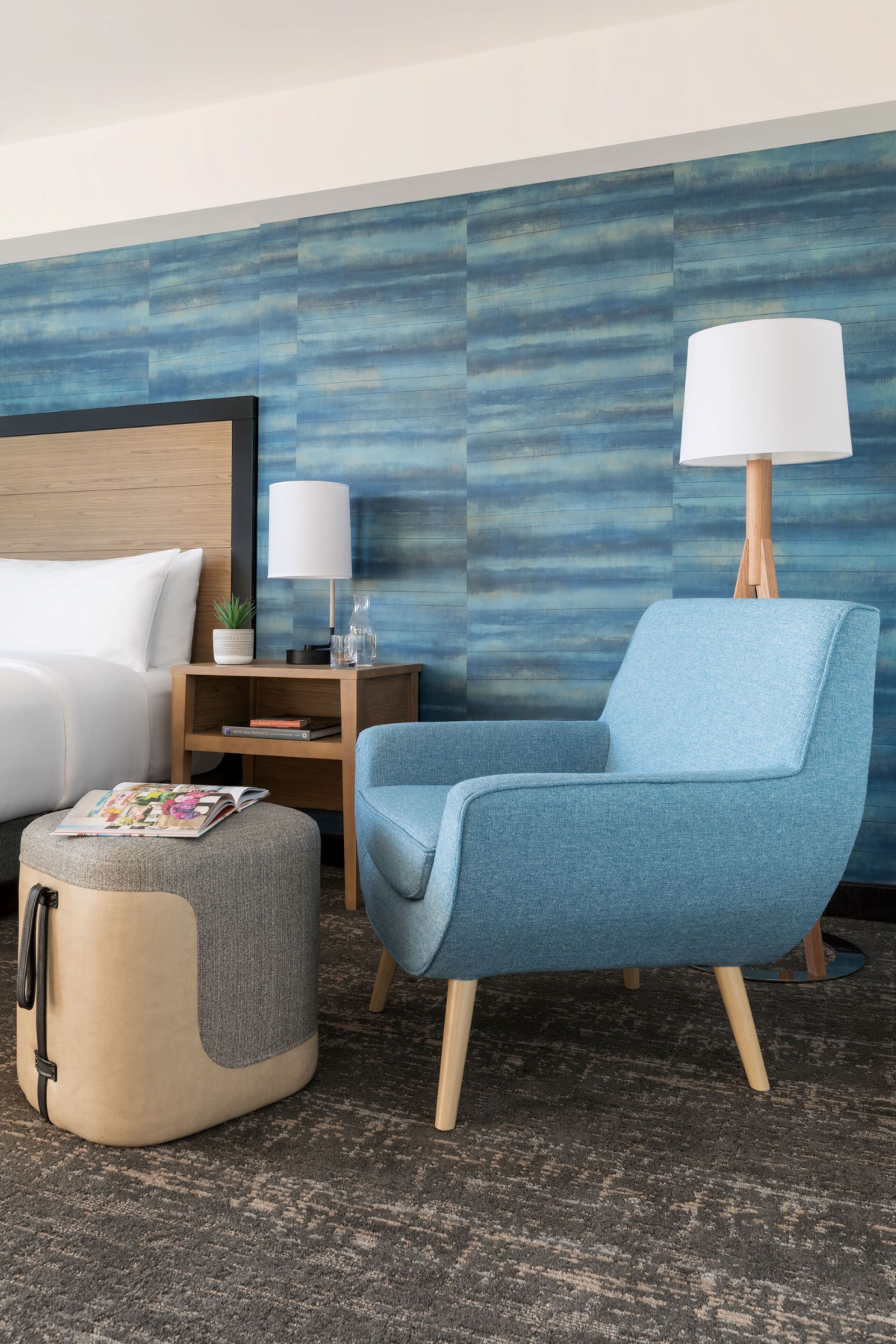 A modern hotel room with a bed, a blue armchair, a small ottoman, a nightstand with a lamp, and a wall with a blue abstract pattern.
