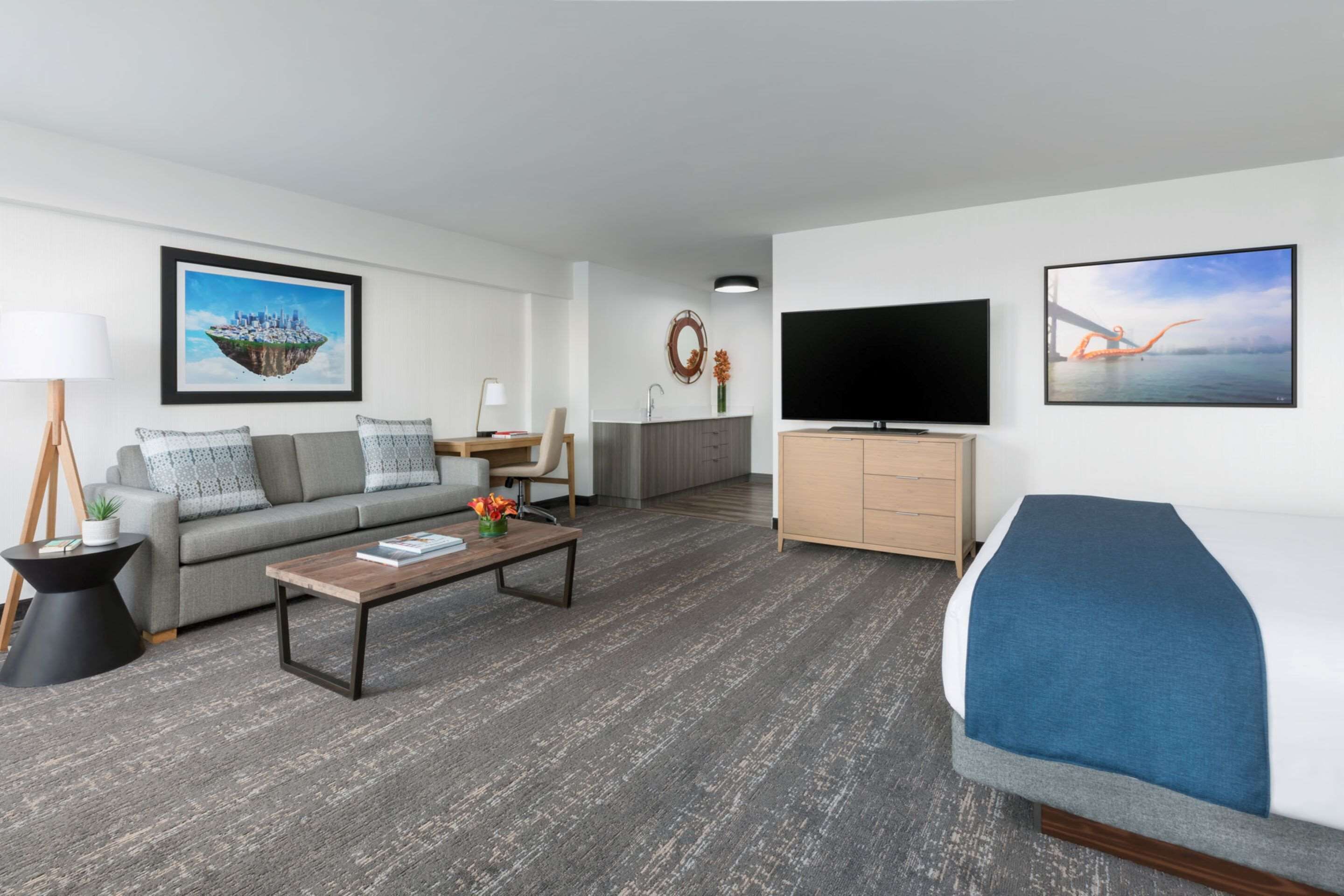A modern hotel room with a sofa, a coffee table, a TV, a bed, and a small kitchenette.