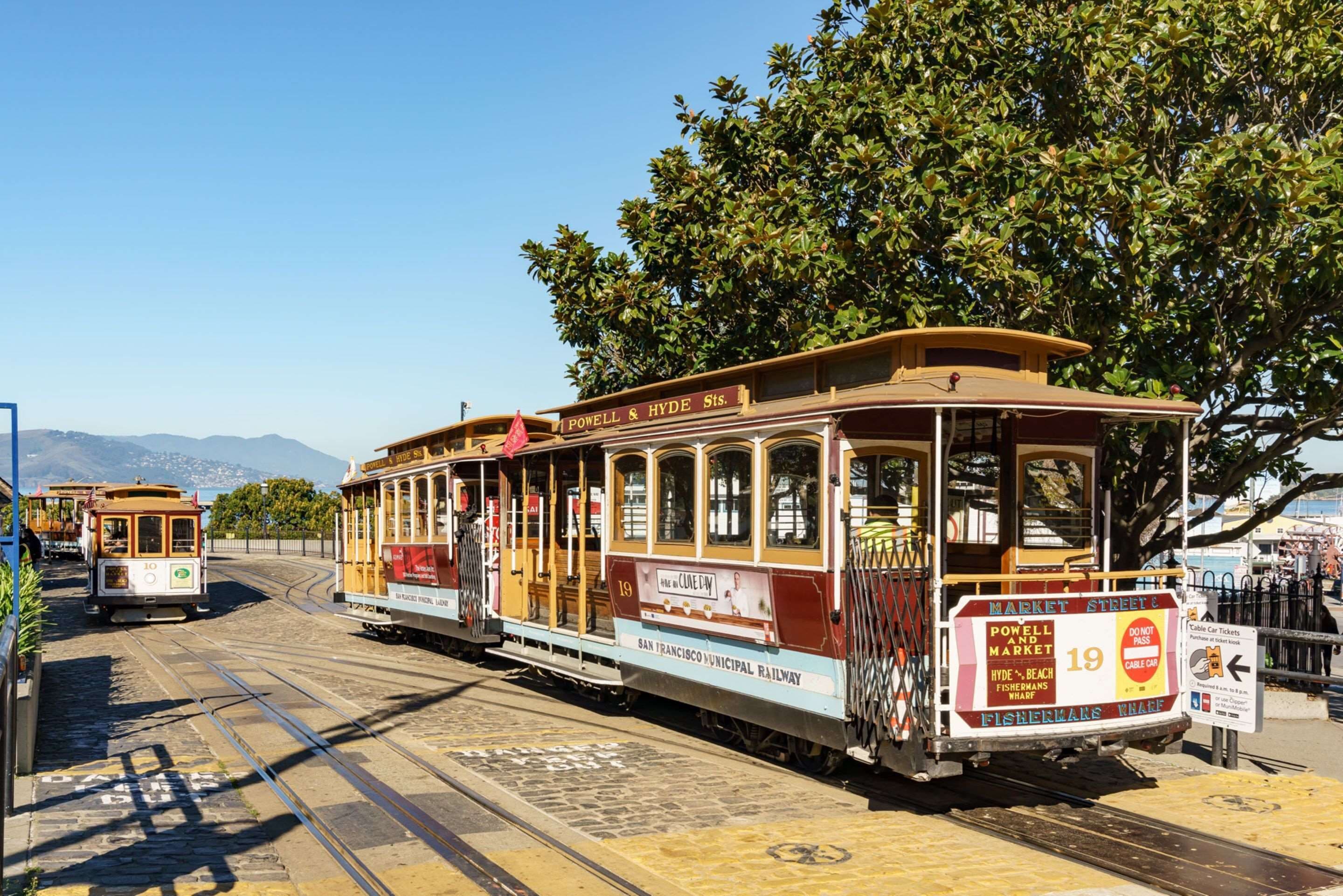 Un tranvía antiguo con letreros que dicen "Powell & Hyde Sts." y "San Francisco Municipal Railway", circulando sobre rieles con montañas al fondo.